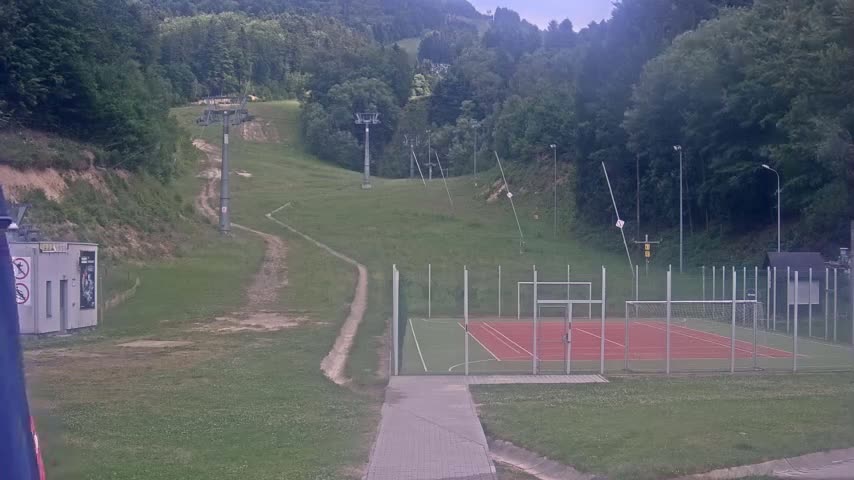 A paved walkway leads to an outdoor sports court, bordered by a grassy hill with ski lifts and a forested hillside in the background under an overcast sky.