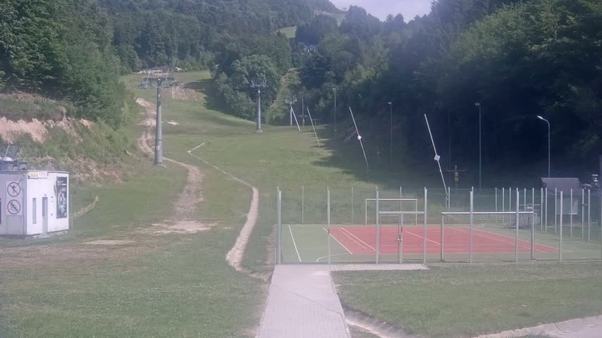 A paved walkway leads to an enclosed multi-use games area situated below a grassy hillside with ski lifts and a forested background on a sunny day.