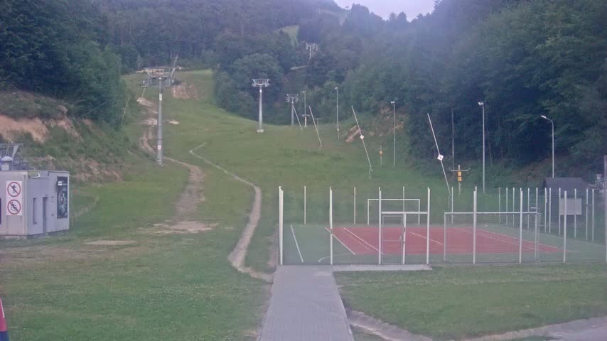 A paved walkway leads to an enclosed sports court next to a grassy hillside with several ski lift towers visible in the background on an overcast day.