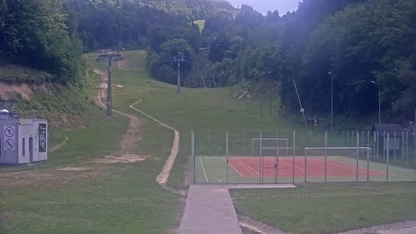 A multi-use games area sits below a grassy hillside with a chairlift and trails visible on an overcast day.