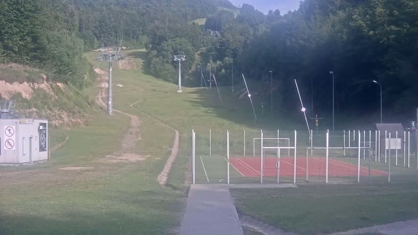 A grassy hillside with ski lift infrastructure, a fenced-in sports court, and a small building is visible on a partly sunny day.