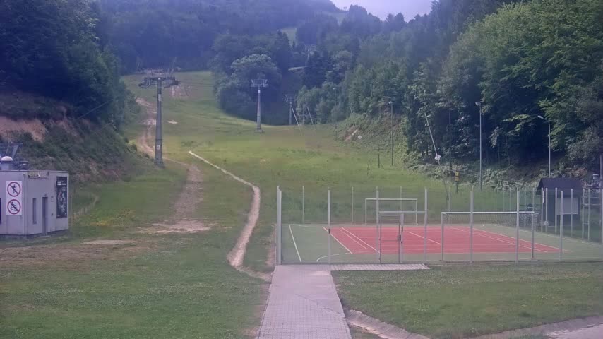 A paved walkway leads to an enclosed sports court, situated below a grassy hillside with two ski lifts and a dense forest in the background on an overcast day.