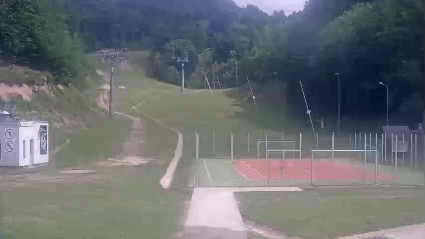 A paved path leads to an outdoor sports court surrounded by a fence, situated on a grassy area with a ski lift and wooded hills in the background under an overcast sky.