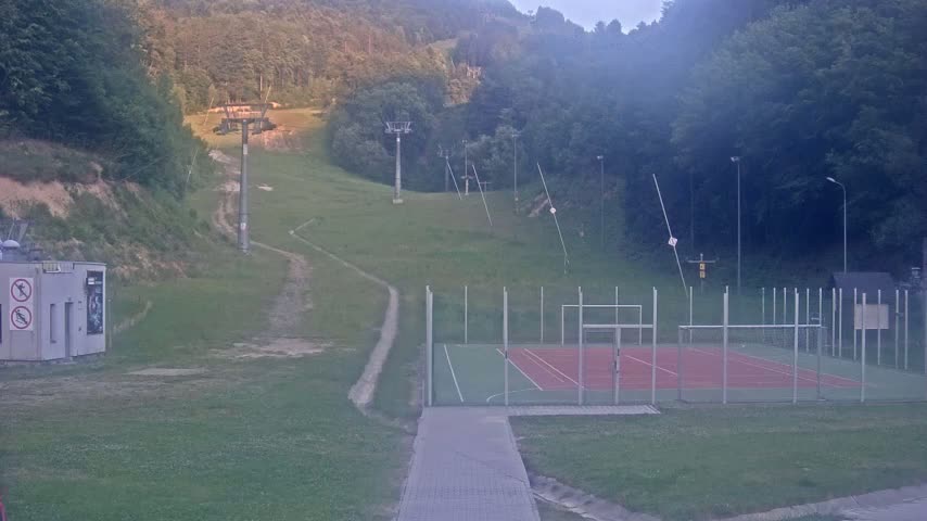 A paved walkway leads to a multi-purpose sports court enclosed by a fence, situated below a grassy hillside with ski lift infrastructure and a wooded backdrop under an overcast sky.