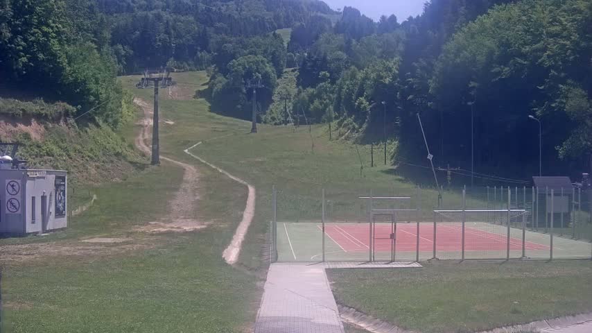 A paved walkway leads to an outdoor tennis court, situated below a grassy hillside with chairlifts and lush green trees under a sunny sky.