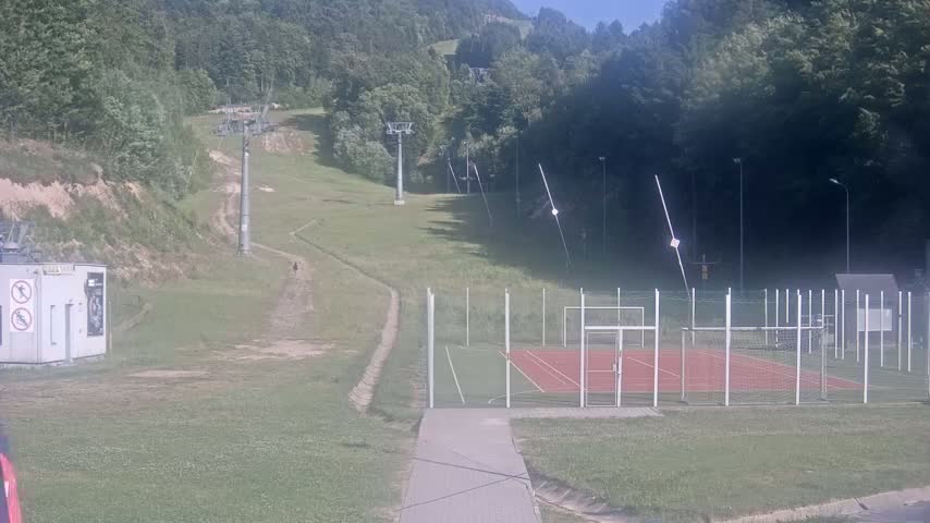 A grassy hill with ski lifts in the background, a small enclosed sports court in the foreground, and sunny weather.