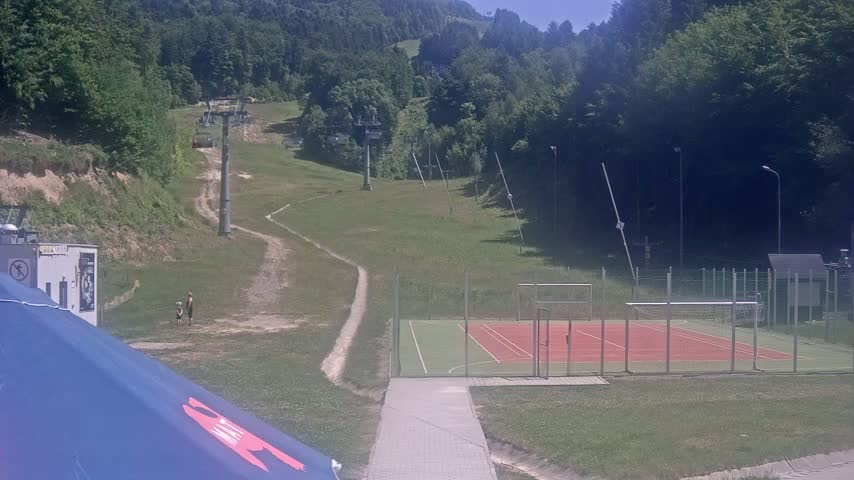 A grassy hillside with a ski lift and a multi-purpose sports court is visible on a sunny day.