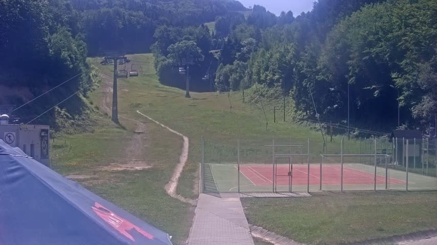 A grassy hillside with a chairlift, a paved walkway leading to an enclosed tennis/basketball court, is visible on a sunny day.