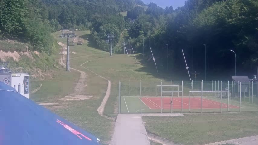 A grassy hillside with inactive ski lifts, a multi-purpose sports court, and a paved walkway, all under a sunny sky.