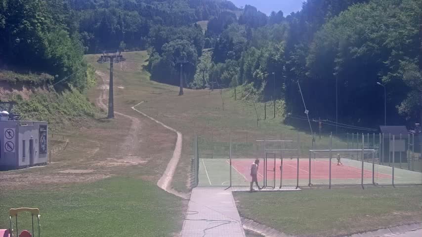 A grassy hillside with a ski lift, a paved pathway leading to an enclosed tennis court with two people, and a small building in the distance on a sunny day.