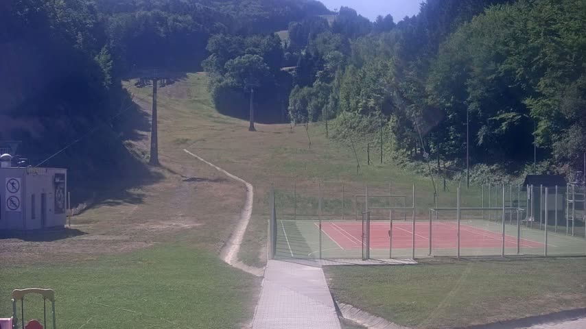 A paved walkway leads to an enclosed tennis court situated below a grassy hillside with a ski lift and surrounding woods on a sunny day.