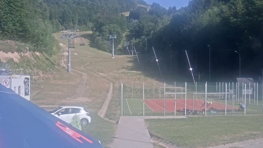 A grassy hillside with several ski lifts, a small car parked nearby, and a tennis court with a few people visible on a sunny day.