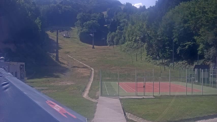 A paved walkway leads to a tennis court enclosed by a fence, situated on a grassy area below a ski lift ascending a hillside covered in trees under a sunny sky.