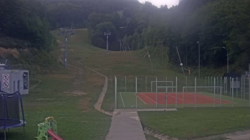 A paved walkway leads to a tennis court enclosed by a fence, situated on a grassy area below a ski lift ascending a hillside covered in trees under a sunny sky.