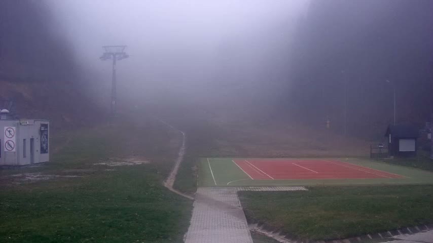 A red and green sports court and a paved path are visible between two small buildings, while a ski lift tower and tree-covered hills disappear into a dense fog on an overcast day.