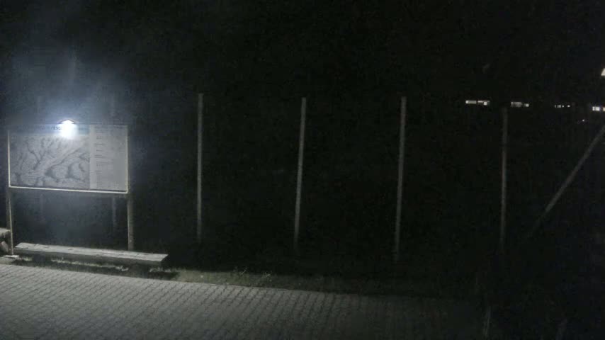 A nighttime outdoor scene shows a lit-up informational sign next to a low bench in front of a dark fence.