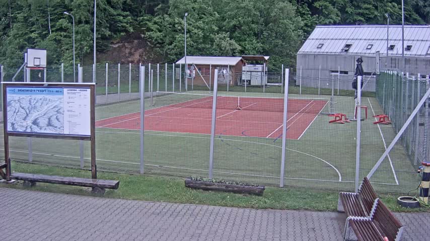 An outdoor tennis and basketball court with a chain-link fence, surrounded by trees and a building, on an overcast day.