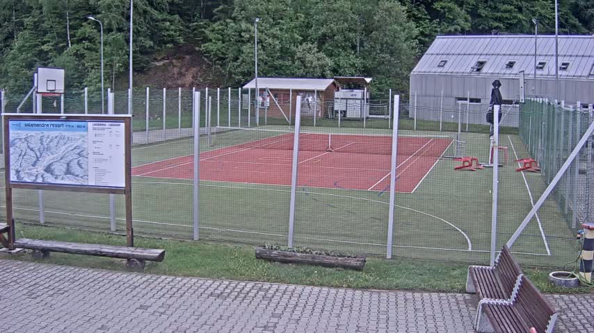 An outdoor multi-use sports court with a red surface, surrounded by a chain-link fence, is situated next to a small building, under an overcast sky.
