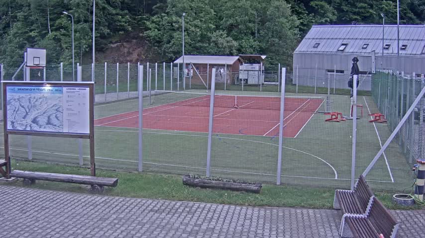 An outdoor tennis and basketball court, enclosed by a chain-link fence, is situated near a small building and a wooded area on an overcast day.