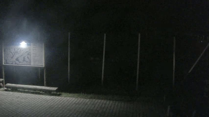A nighttime scene shows a lit-up informational sign with a map and a bench in front of a dark fence.