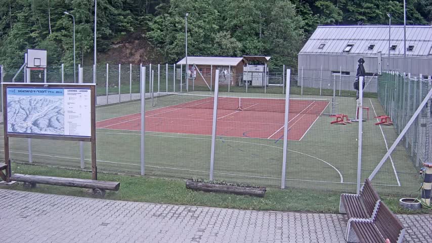 An outdoor multi-purpose sports court with a red surface, surrounded by a chain-link fence, sits under an overcast sky.