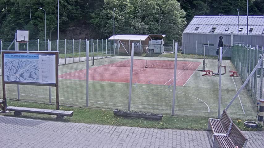 An outdoor multi-use court with a red surface is enclosed by a chain-link fence, with a basketball hoop and other sports equipment visible.