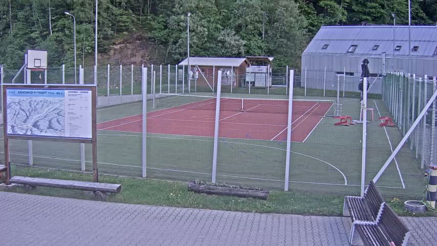 An outdoor multi-purpose court with a red surface, surrounded by a chain-link fence, sits adjacent to a small building and a basketball hoop.
