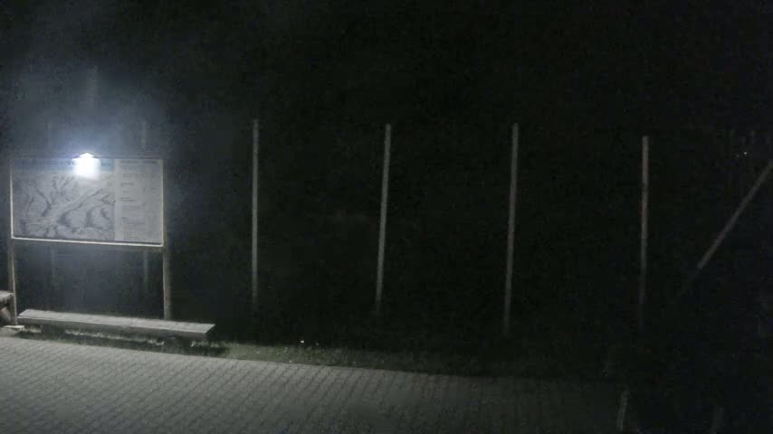 A nighttime scene shows a lighted informational sign with a bench in front of a dark fence.