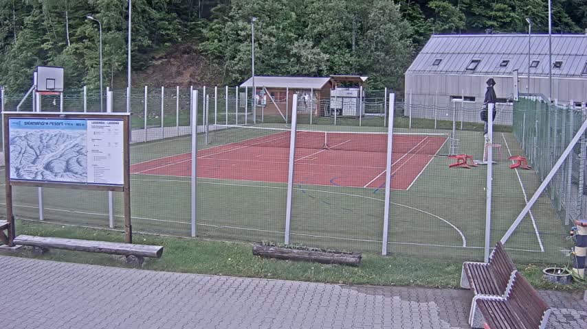 An outdoor multi-use sports court with a tennis court and basketball court, surrounded by a chain-link fence, on an overcast day.