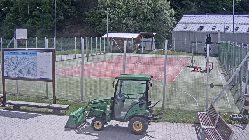 A green tractor with a bucket attachment sits beside an outdoor multi-use sports court enclosed by a fence on a sunny day.