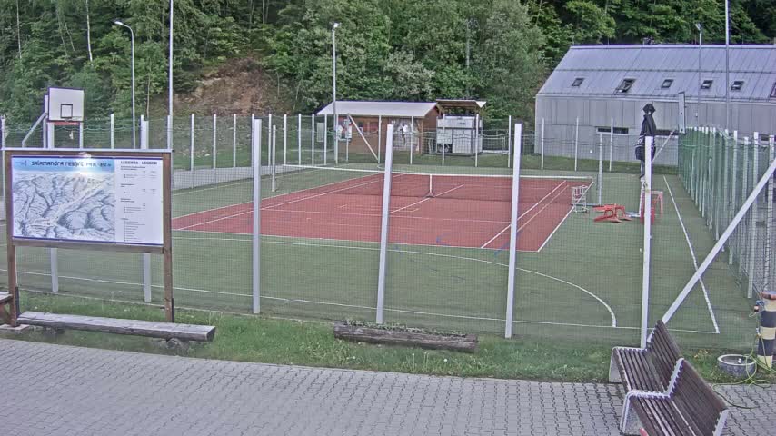 An outdoor tennis court and basketball court, enclosed by chain link fencing, are situated near a small building and surrounded by trees under a partly cloudy sky.