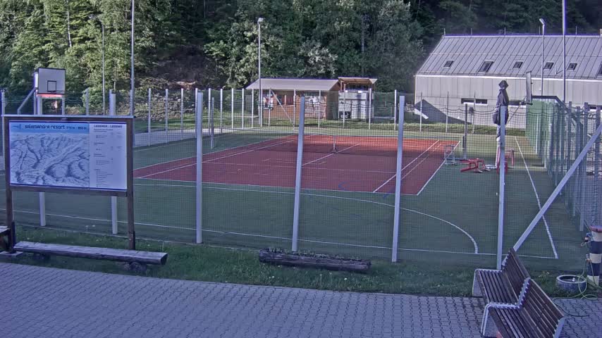 An outdoor multi-purpose sports court with a basketball hoop and tennis markings is enclosed by a fence, situated near a building and wooded area under overcast skies.