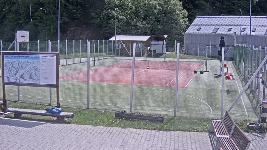 A tennis and basketball court surrounded by a chain-link fence is situated outdoors on a partly sunny day, near a building and some benches.