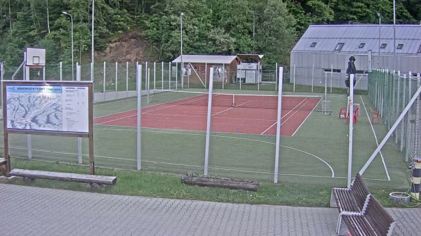 An outdoor multi-use sports court with a red tennis court surface and green surrounding area, enclosed by a fence, is visible under overcast skies.