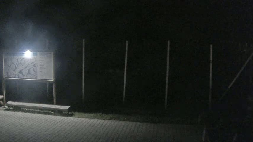 A nighttime outdoor scene shows a lit map on a signpost with a bench in front of a dark fence.