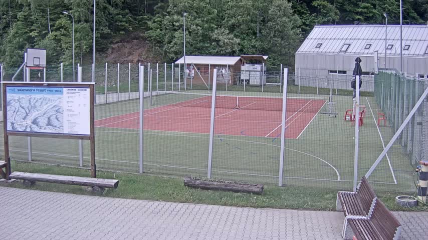 An outdoor multi-purpose sports court with a tennis court and basketball hoop is surrounded by a chain-link fence on an overcast day.