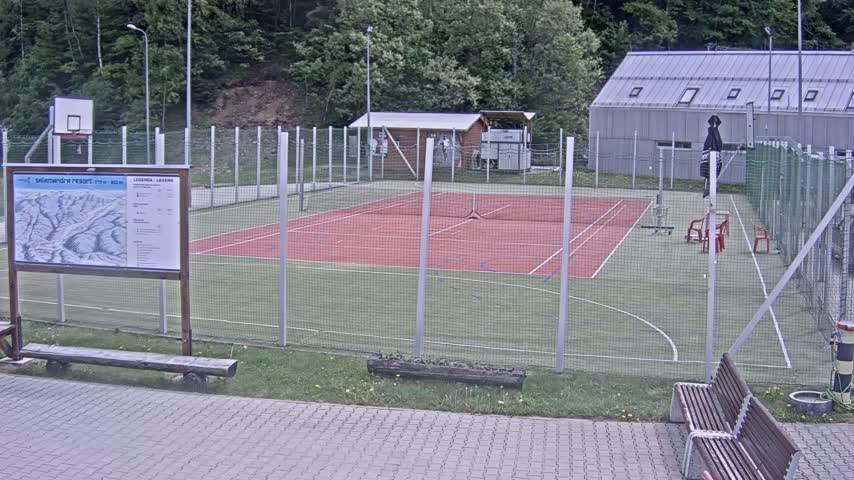 An outdoor multi-purpose court, enclosed by a chain-link fence, features a red tennis/basketball court and a basketball hoop; the area is partially shaded and surrounded by trees and a building.