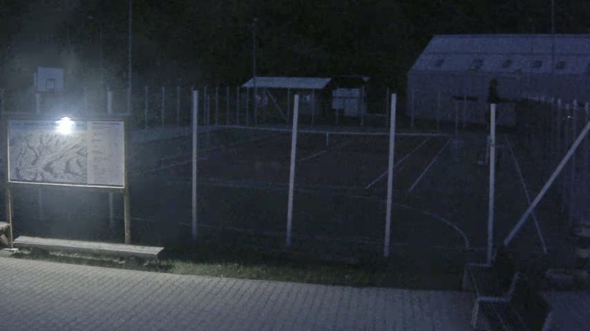An outdoor, nighttime scene shows an enclosed tennis court, a small building, and a map illuminated by a light.