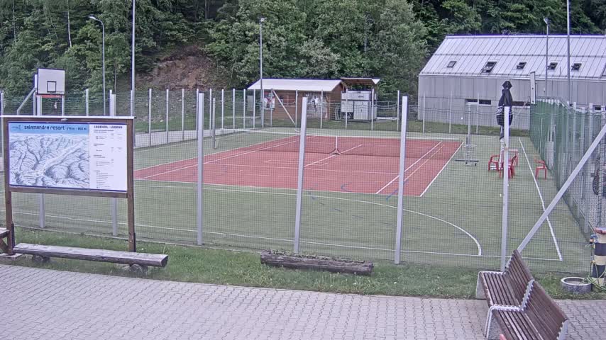 An outdoor multi-purpose court with a red tennis court surface and a basketball hoop is enclosed by a wire fence on a partly cloudy day.