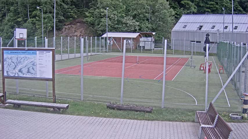 An outdoor multi-use sports court with a red surface, surrounded by a chain-link fence, is situated next to a wooded area on an overcast day.