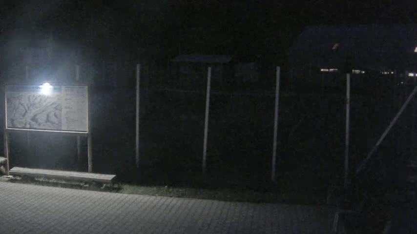 A lighted map is mounted on a post next to a bench at night, in front of a dark fence and building.