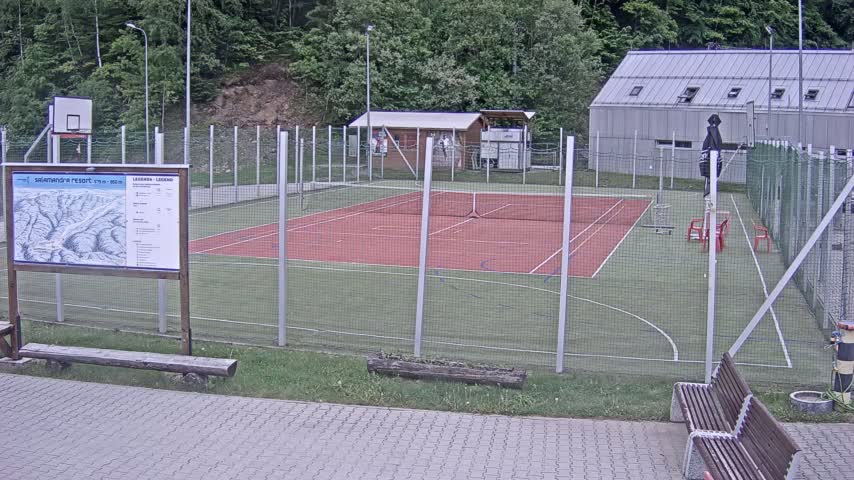 An outdoor multi-use court with a red surface and green surrounding area is enclosed by a chain-link fence, under an overcast sky.