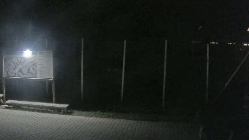 A dimly lit nighttime scene shows a map on a signpost in front of a dark fence and paved walkway.