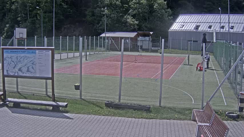 An outdoor tennis and basketball court with a chain-link fence, surrounded by green grass and trees on a sunny day.