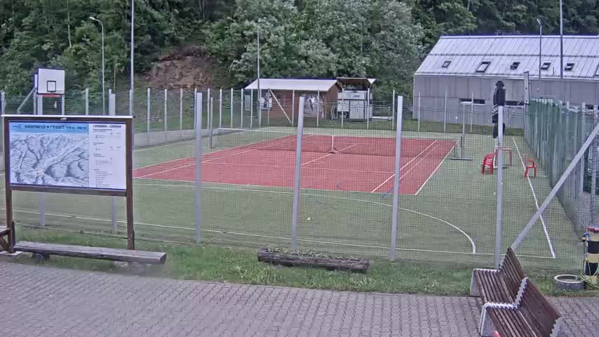 An outdoor tennis court and basketball court are enclosed by a chain-link fence, with a small building and trees in the background under overcast skies.