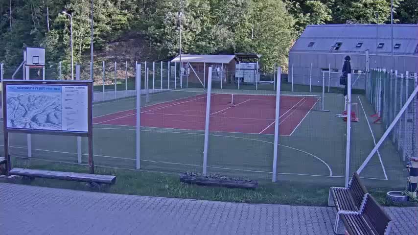 An outdoor multi-purpose court with a red surface, surrounded by a chain-link fence, sits next to a small building under a partly cloudy sky.