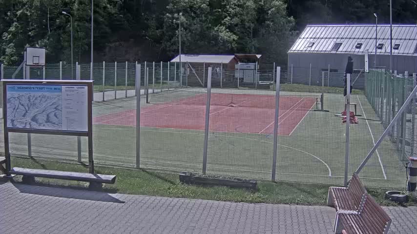 An outdoor multi-purpose sports court with a basketball hoop and benches is surrounded by a chain-link fence on a partly sunny day.