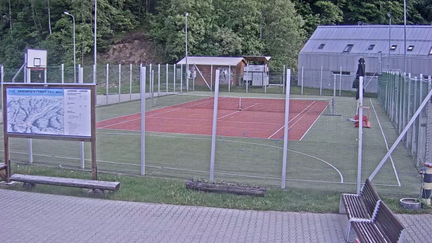 An outdoor multi-purpose sports court with a red surface and green surrounding turf is enclosed by a chain link fence, with a building and trees visible in the background under overcast skies.