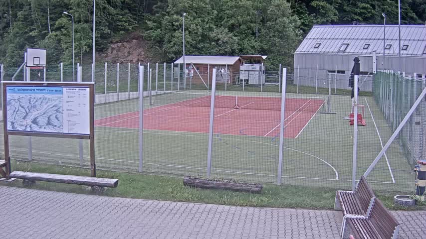An outdoor multi-purpose court with a red surface and green surrounding area, enclosed by a wire fence, is situated near a small building and forested backdrop in overcast conditions.