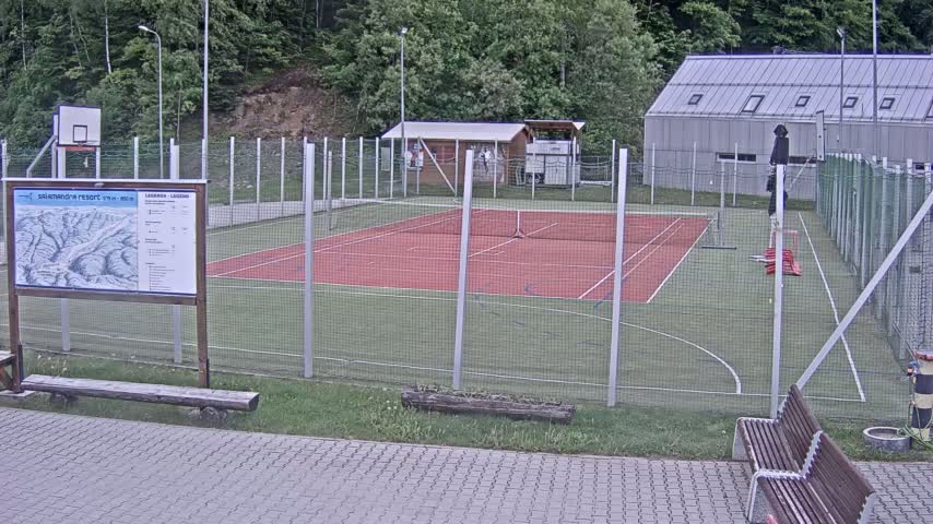 An outdoor multi-use sports court with a red surface, surrounded by a chain-link fence, sits beside a small building and a basketball hoop, all under an overcast sky.